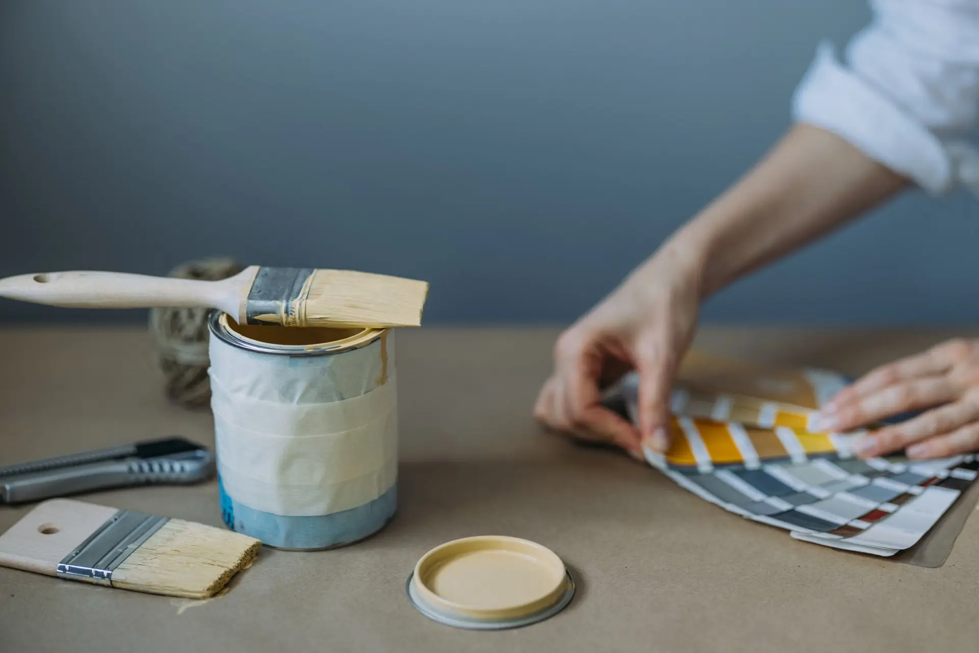 Hands flipping through a paint colour fan deck beside an open paint tin and brushes